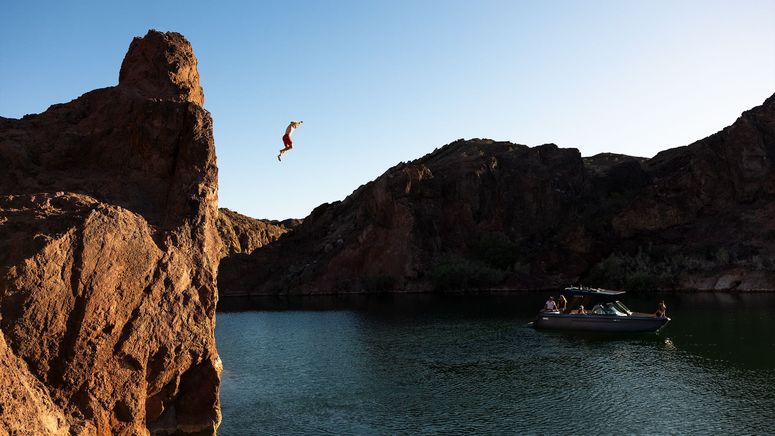 A photograph of a man jumping off of a cliff into a lake with a boat waiting for him.